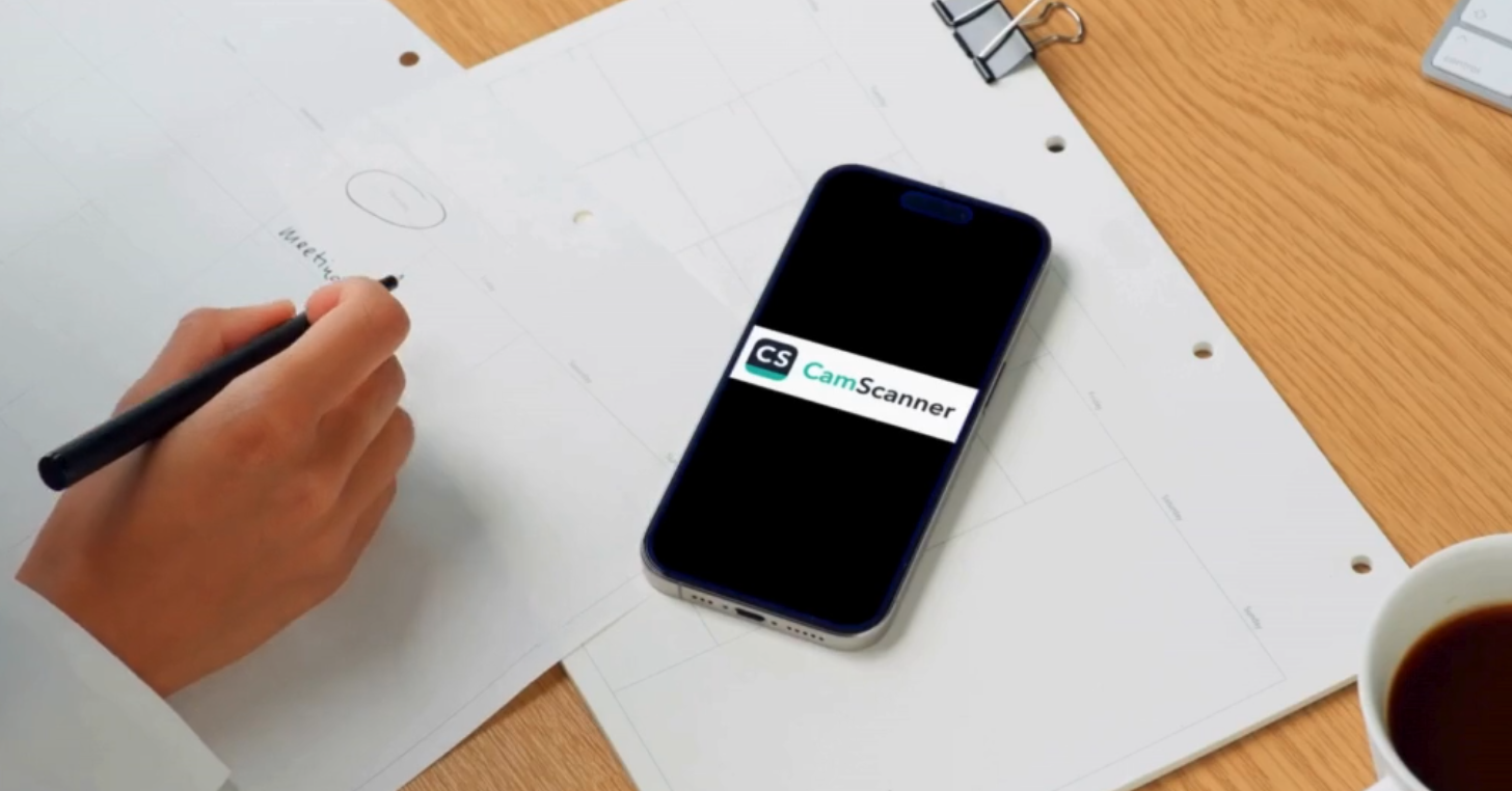 رجل يجلس أمام طاولة عليها أوراق وهاتف ذكي يعرض شعار تطبيق CamScanner المستخدم لتصوير الوثائق وتحويلها إلى ملفات رقمية